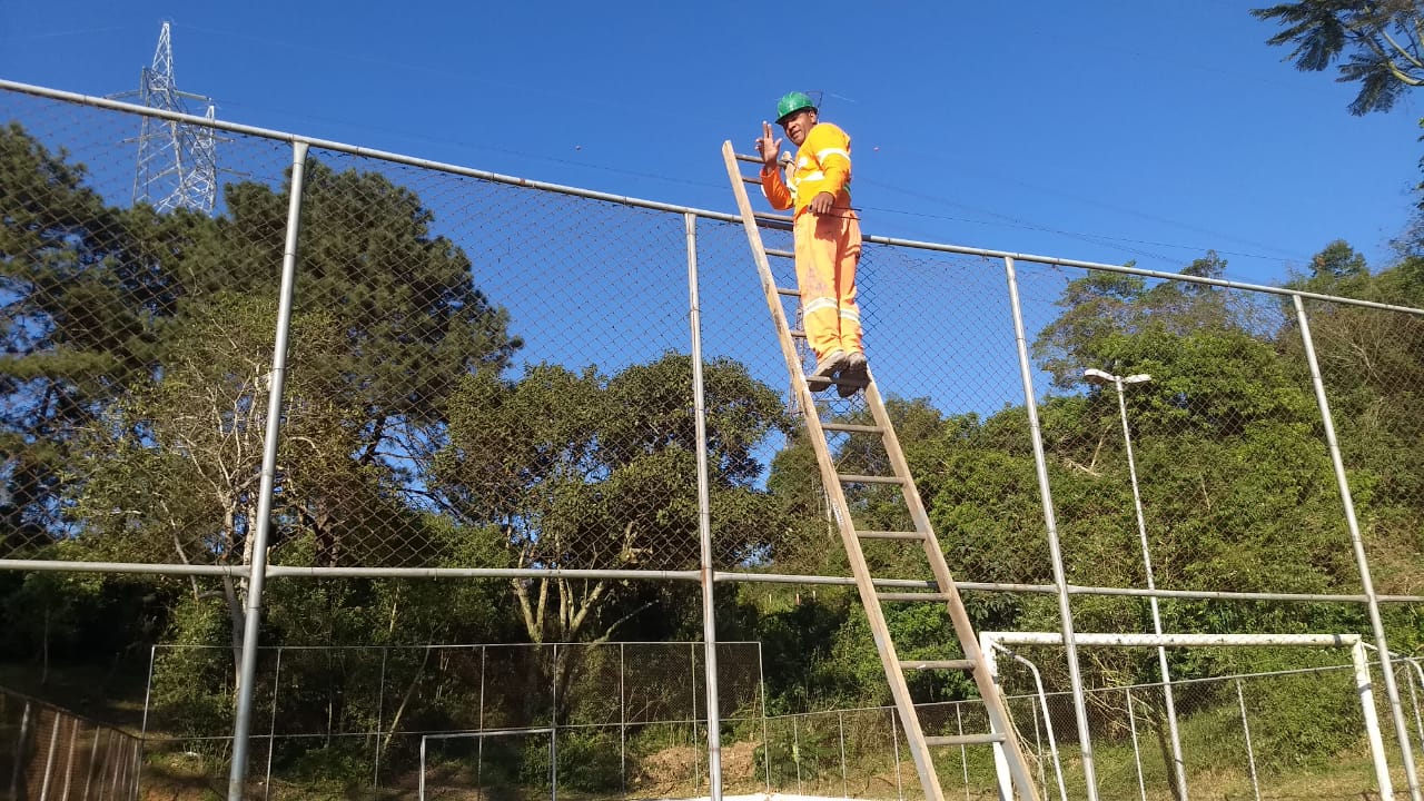 #PraCegoVer - Trabalhador da Subprefeitura, em cima de uma escada, conserta o alambrado da quadra no   Bororé. Ao fundo, há farta vegetação.