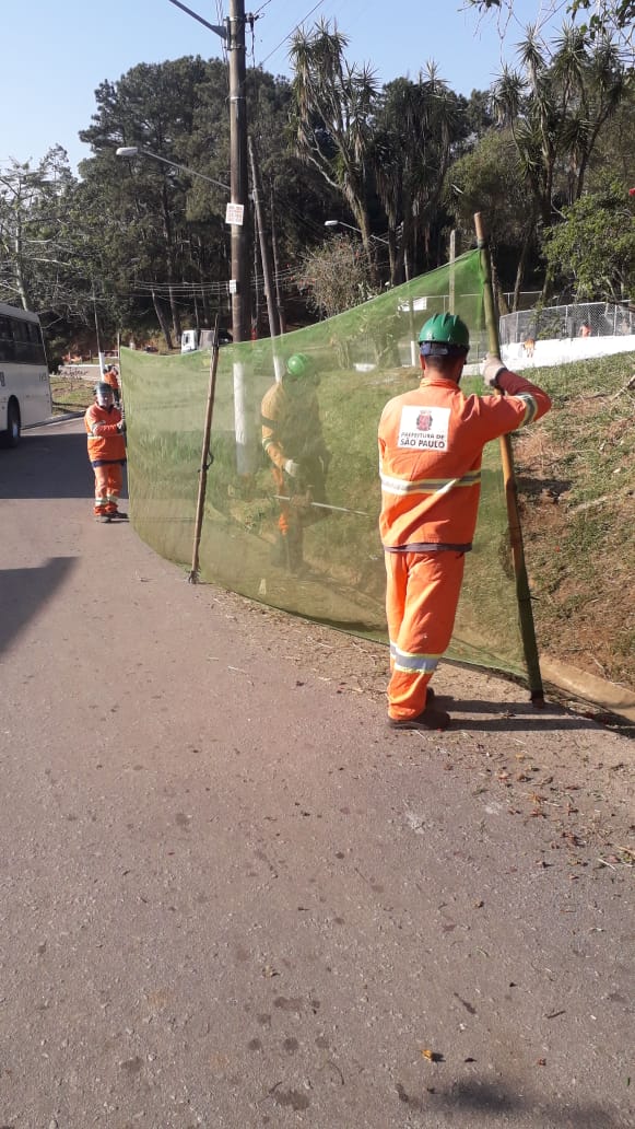 #PraCegoVer - Dois trabalhadores da Subprefeitura seguram tela de proteção enquanto um terceiro corta a grama à beira da estrada que atravessa a Ilha do Bororé.