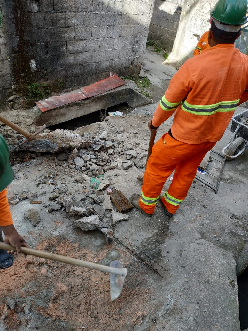 #PraCegoVer - Dois trabalhadores da Subprefeitura abre a via para expor a galeria de águas pluviais (GAP). Há fragmentos de concreto espalhados pelo chão.