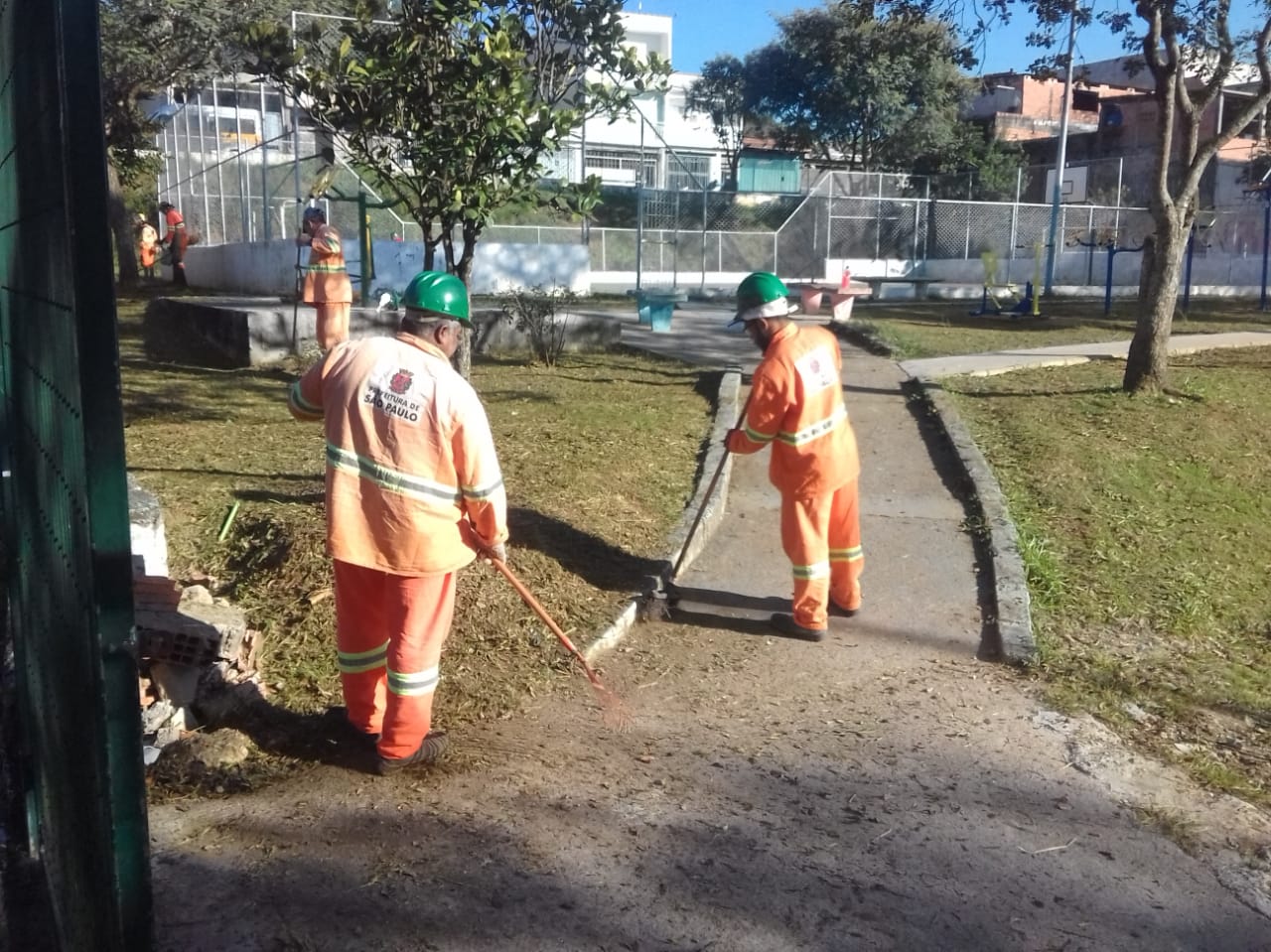 #PraCegoVer - Cinco trabalhadores da Subprefeitura cortam grama e limpam os passeios da praça. Há uma quadra fechada por alambrado ao fundo. E uma árvore à direita.