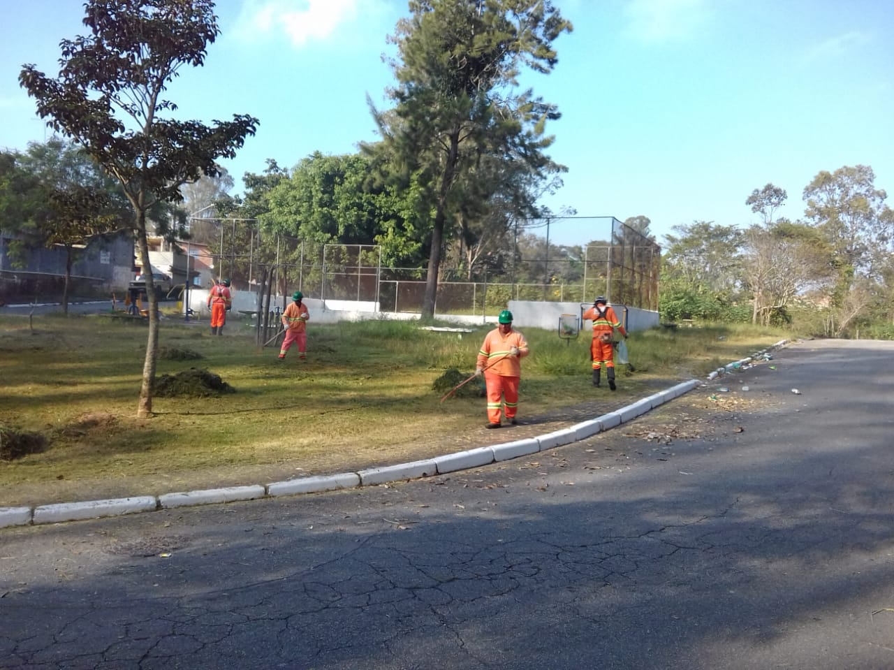 #PraCegoVer - Quatro trabalhadores da Subprefeitura cortam grama e juntam a grama já cortada em uma curva da via. Ao fundo, há uma quadra de esportes com alambrado, cercada por árvores.