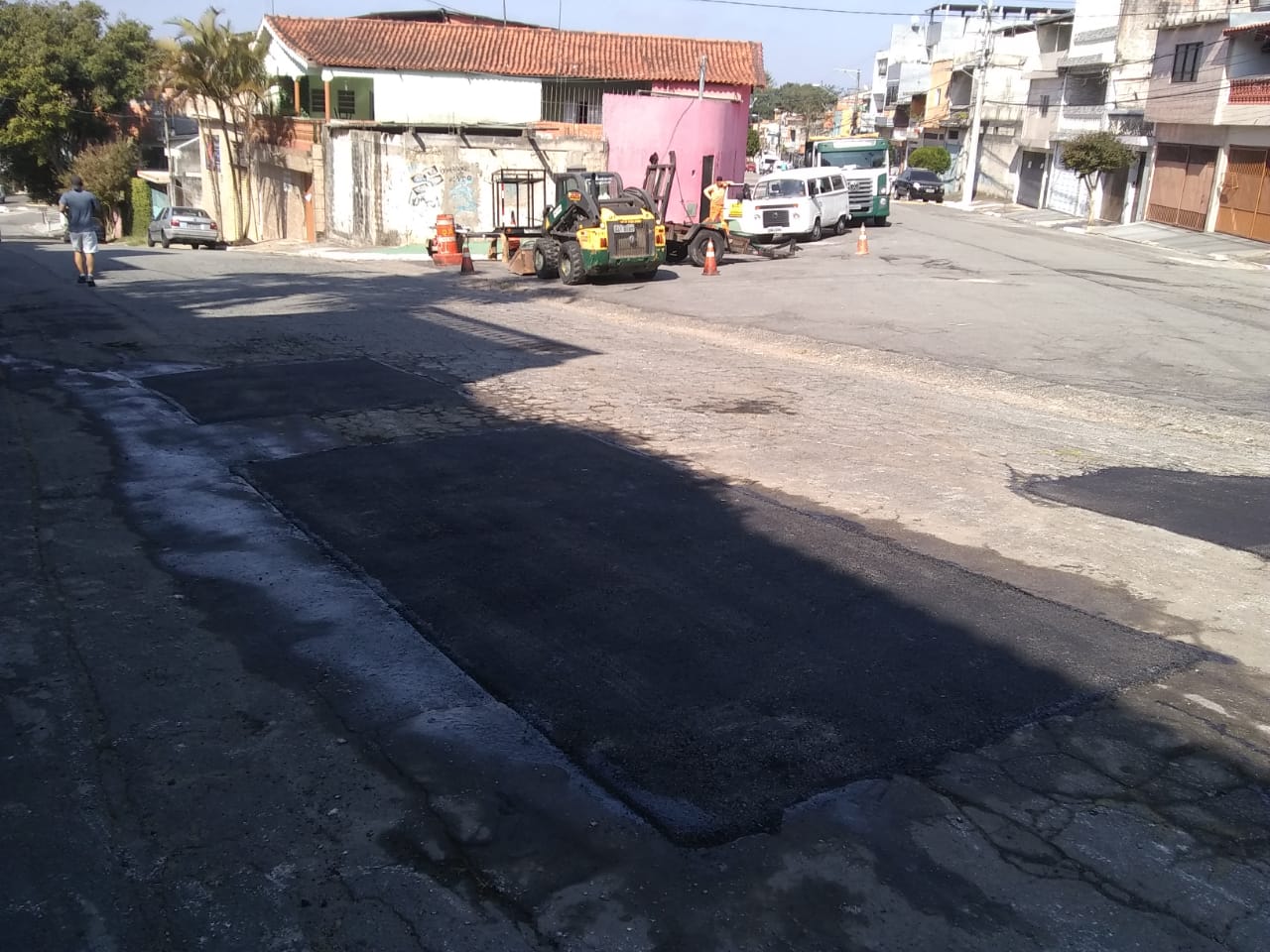 #PraCegoVer - O local fica no entroncamento de duas ruas. O buraco aparece em primeiro plano; na segunda foto, já está tapado. Ao fundo, caminhão da empreiteira que fez o serviço. Mais ao fundo ainda uma Kombi branca.