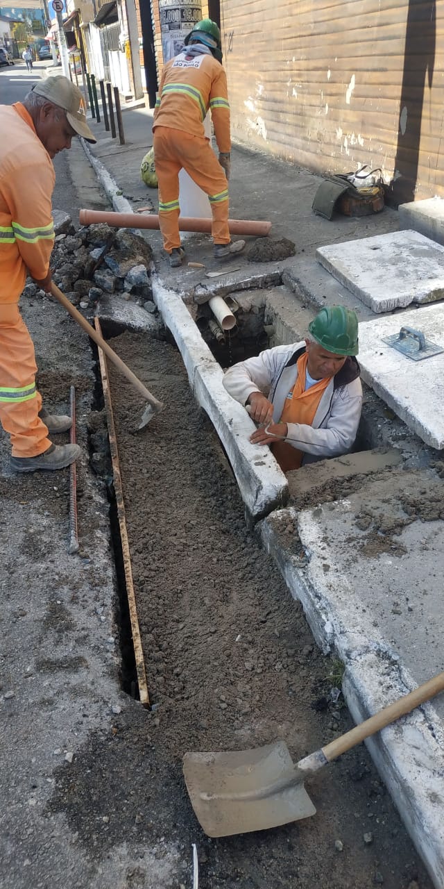 #PraCegoVer - Um trabalhador da Subprefeitura está dentro da boca-de-lobo (BL); as tampas estão afastadas, de lado. Outros dois trabalhadores preparam a sarjeta para concretagem.