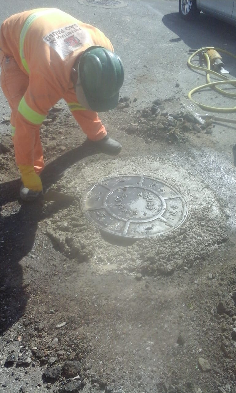 #PraCegoVer - Trabalhador da Subprefeitura faz a finalização em cimento do nivelamento do poço-de-visita (bueiro). Ao fundo há uma britadeira com um cabo amarelo.
