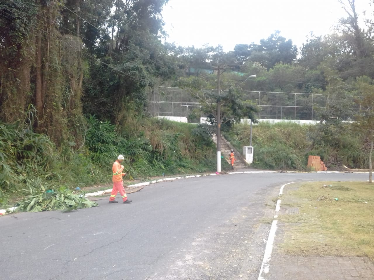 #PraCegoVer - Dois trabalhadores da Subprefeitura — um corta grama; o outro recolhe galhos de árvores. O local fica em uma curva da rua. Ao fundo, vê-se uma quadra de esportes com alambrado. Chega-se à quadra por uma escadaria que sai do nível da rua.