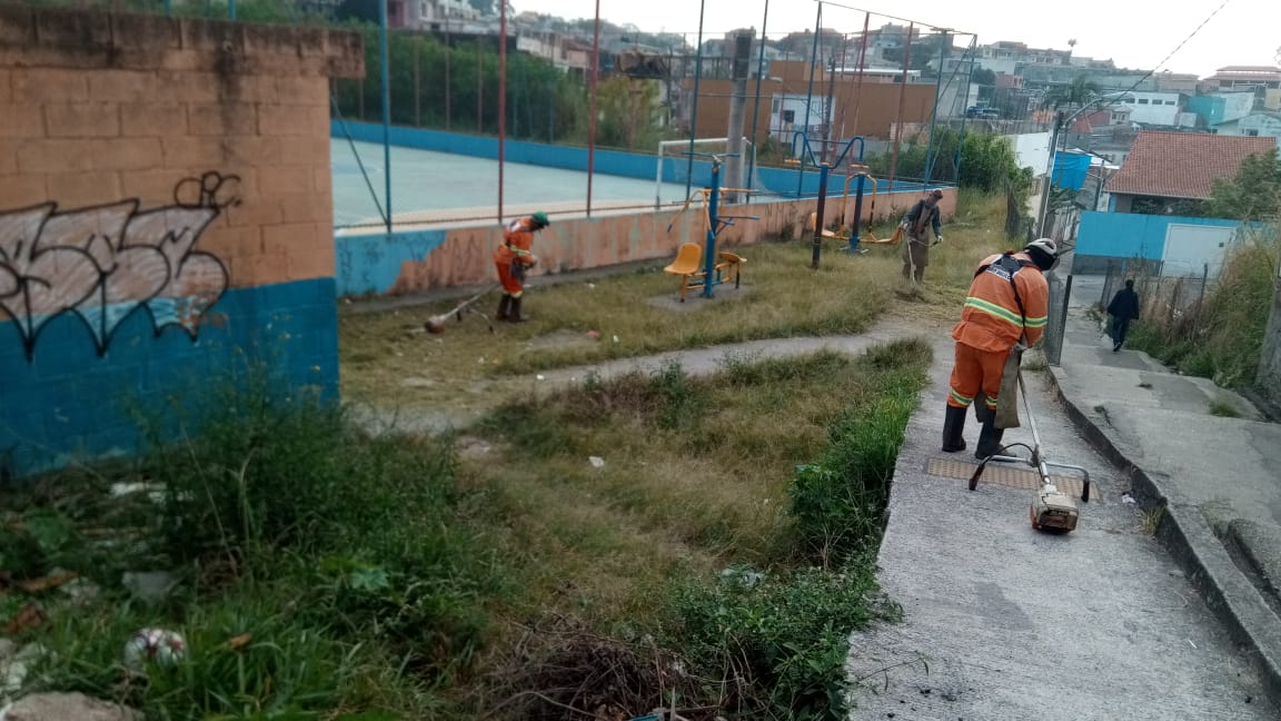 #PraCegoVer - Trabalhadores da Subprefeitura cortam grama em torno de uma quadra na via. Fora da quadra, há aparelhos de ginástica para a Terceira Idade. A quadra é rodeada por casas e por um passeio de concreto.