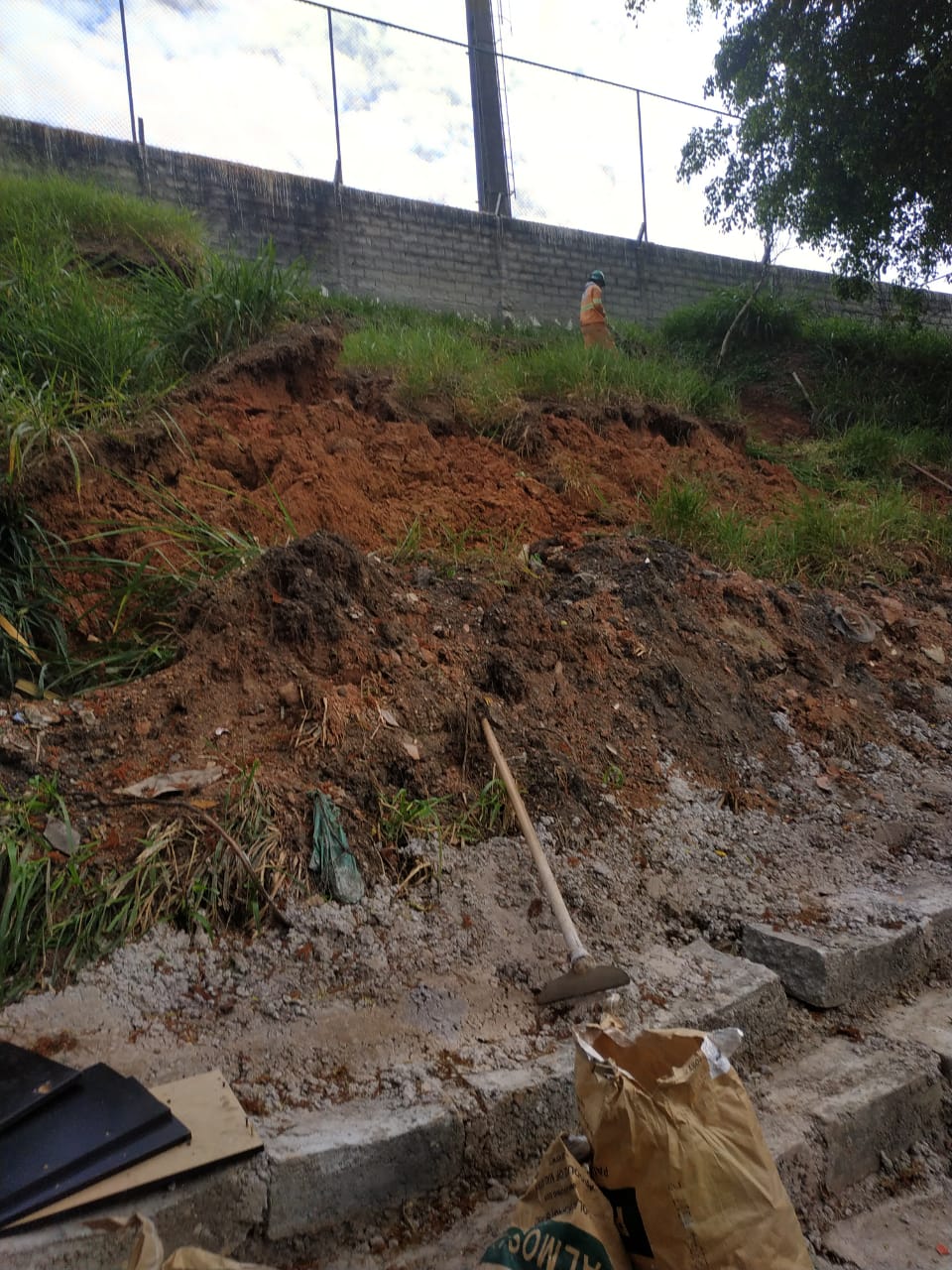 #PraCegoVer - Uma encosta marca o local da reforma da Galeria de Águas Pluviais (GAP). No alto da encosta, há um trabalhador da Subprefeitura. Na parte de baixo, há restos de cimento, uma enxada e sacos de materiais de construção.