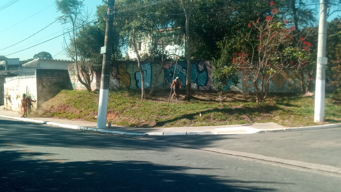 #PraCegoVer - Dois trabalhadores da Subprefeitura cortam grama na praça. O local é uma das quinas da via. Há dois postes no meio e do lado direito. O restante do terreno é pontilhado por árvores.