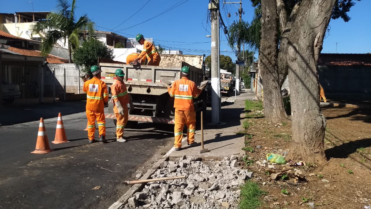 #PraCegoVer - Cinco trabalhadores da Subprefeitura iniciam trabalho de reformar passeio da praça. Parte do passeio já foi fragmentado para facilitar o trabalho. Acompanhando os trabalhadores, há um caminhão da contratada da Subprefeitura. Do lado direito, há duas árvores antigas.