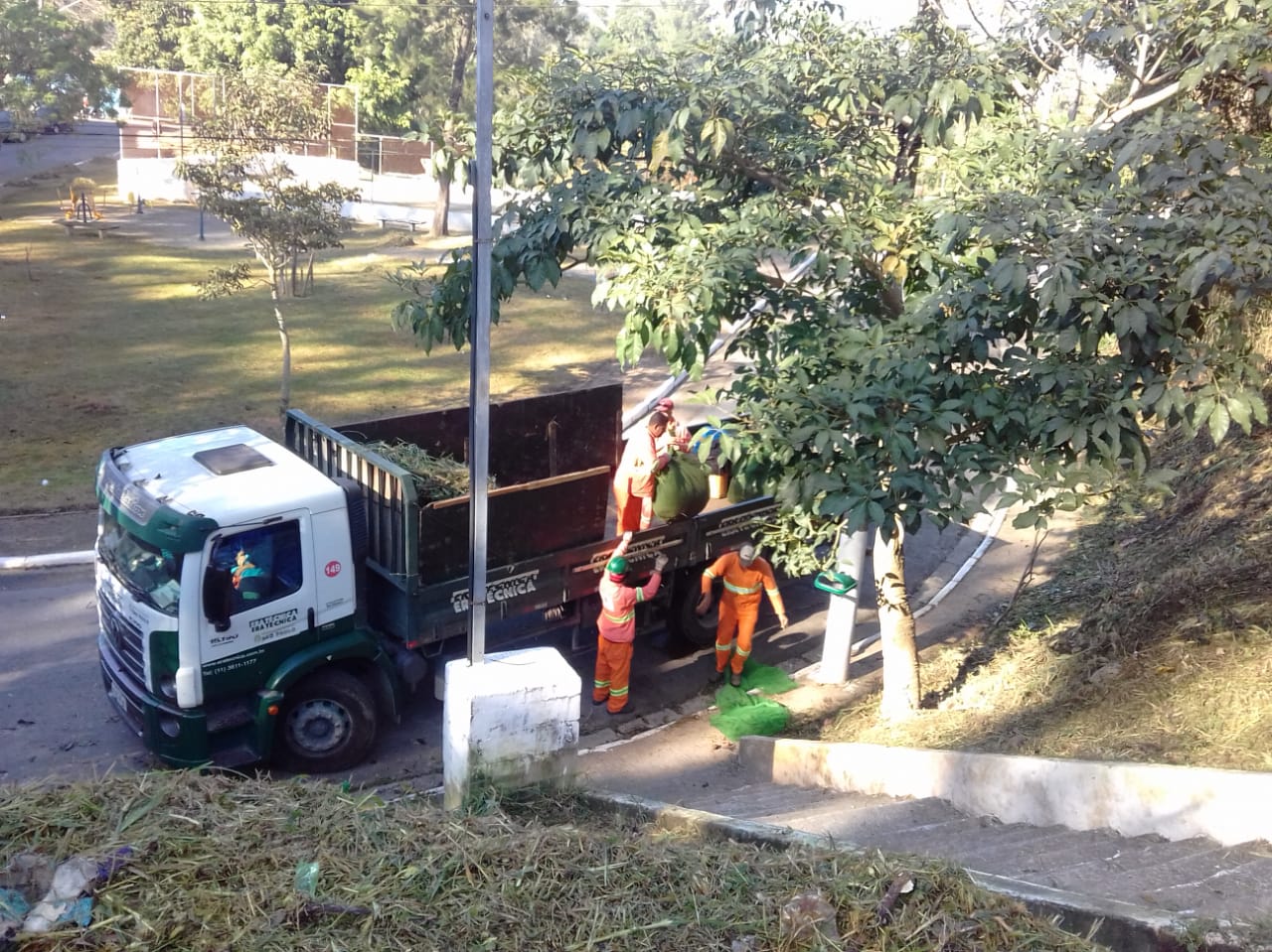 #PraCegoVer - Quatro trabalhadores da Subprefeitura recolhem restos de grama cortados na via. Dois estão no passeio; outros dois, no caminhão que recolhe a grama. Ao fundo, há uma quadra de esportes.