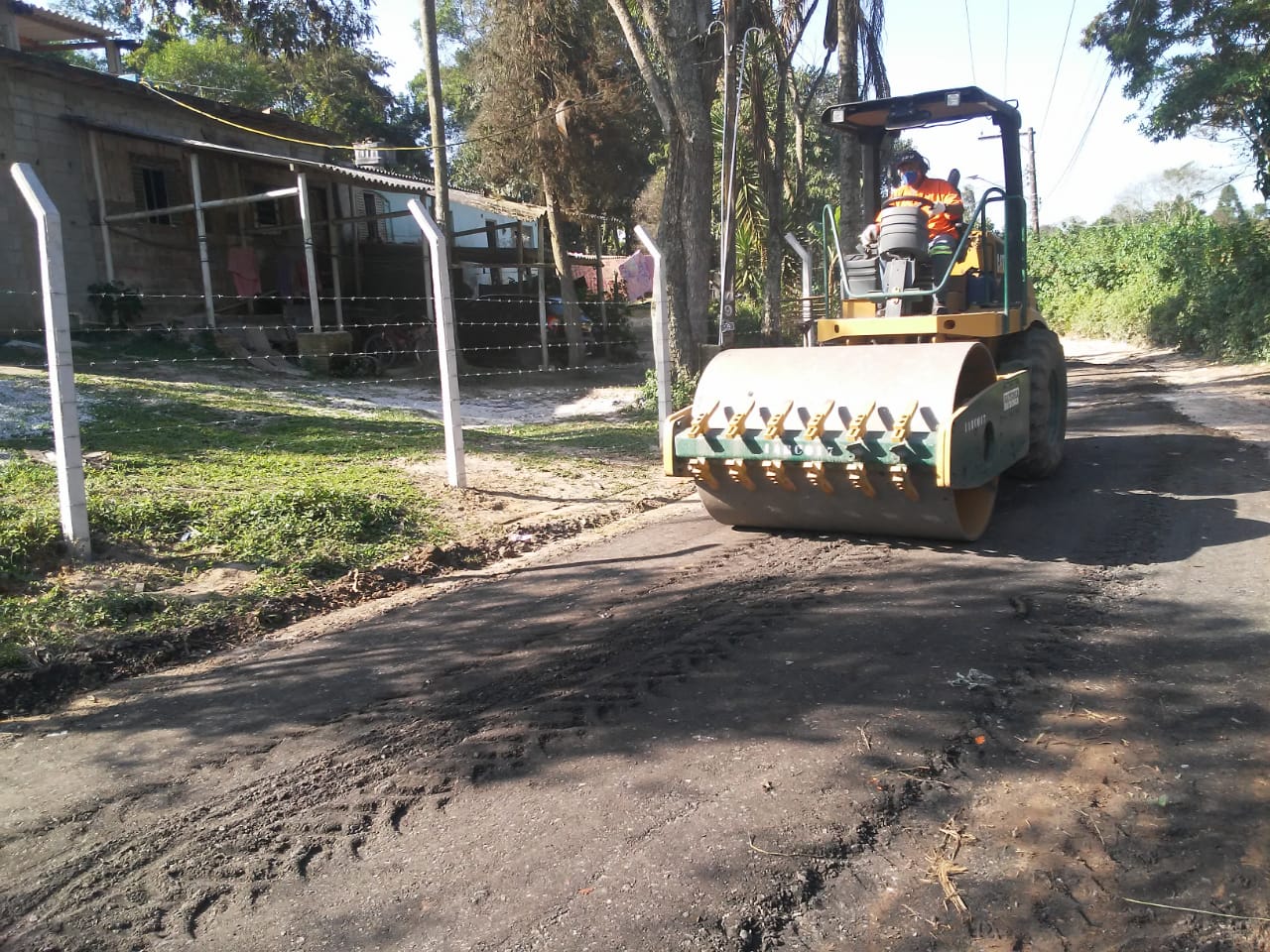 #PraCegoVer - Um rolo compactador passa sobre a terra da via. O local não pode ser asfaltado por ficar em área de mananciais. Do lado esquerdo, há uma construção e uma cerca de arame farpado.