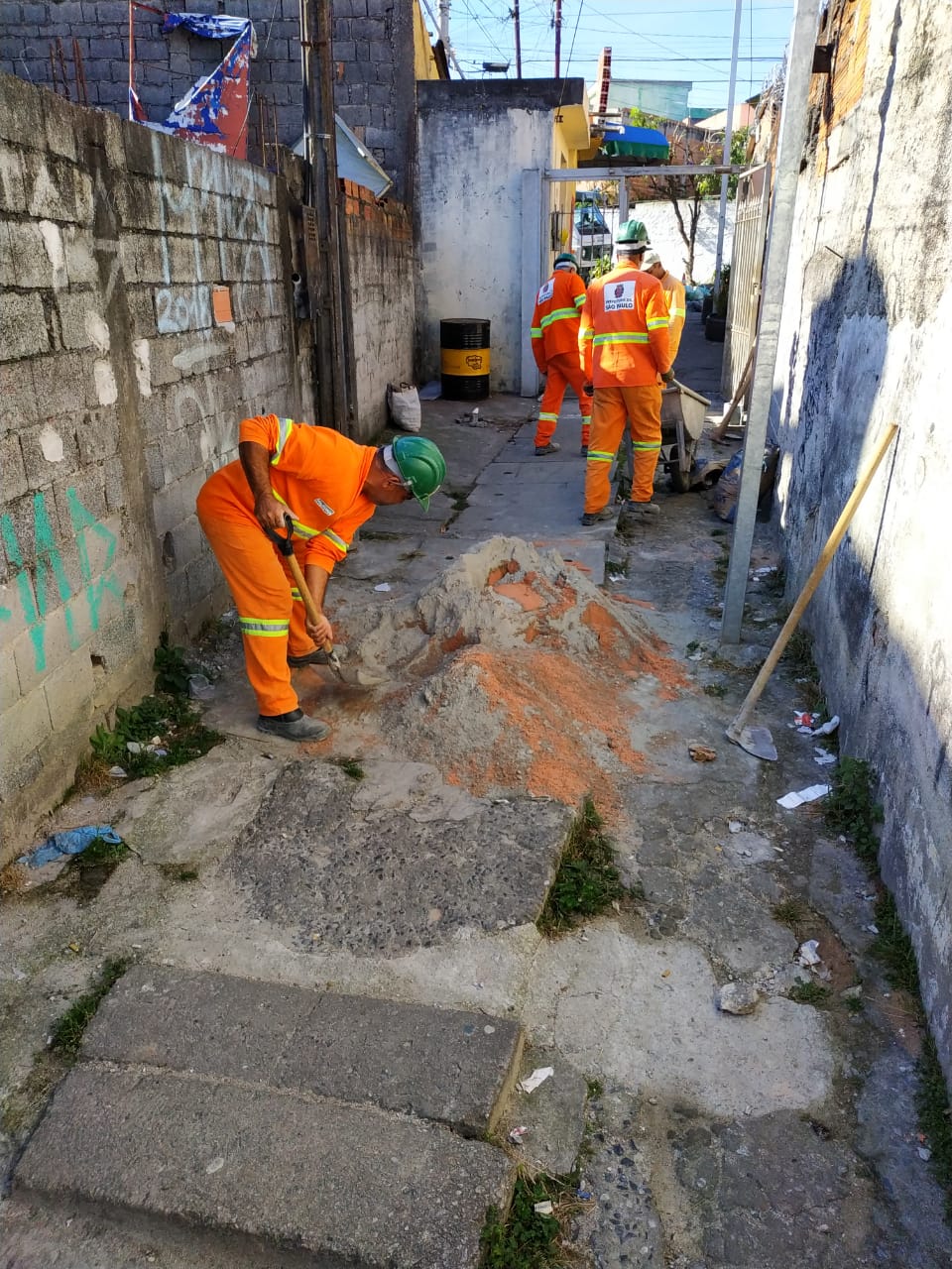 #Pracegover - Em uma viela, três trabalhadores da Subprefeitura preparam cimento para reparar a rede de águas pluviais.
