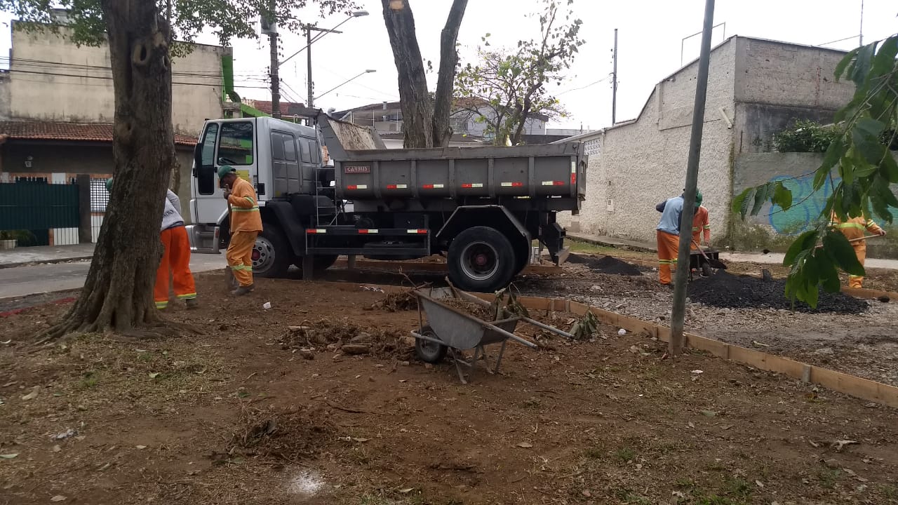 #PraCegoVer - Trabalhadores da Subprefeitura limpam o terreno da praça. Há um caminhão basculante ao fundo. E um carrinho de mão à frente. A praça é cercada por árvores e por casas.