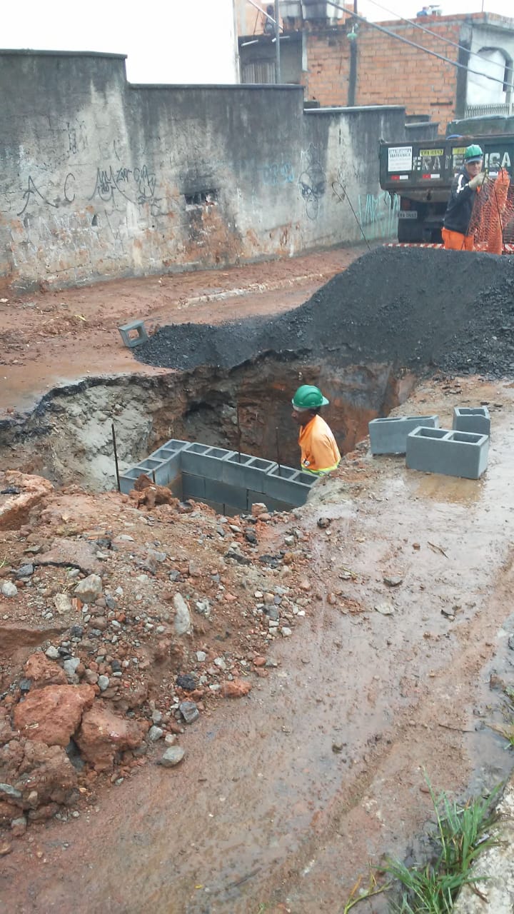 #PraCegoVer - Trabalhador da Subprefeitura está dentro de um buraco cavado na via. Ele assenta blocos de cimento para reconstruir a galeria de águas pluviais. Ao fundo, há outro trabalhador preparando a rede de proteção, e um caminhão da empreiteira contratada.