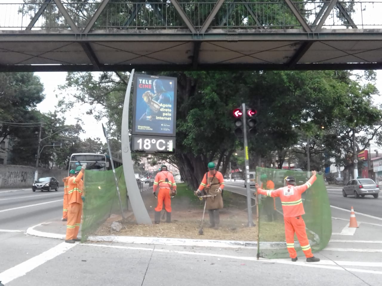 #PraCegoVer - Trabalhadores da Subprefeitura fazem corte de grama na entrada da av. Belmira Marin. Dois cortam a grama. Outros quatro seguram a proteção de tela. Na parte de cima, há uma passarela para pedestres.