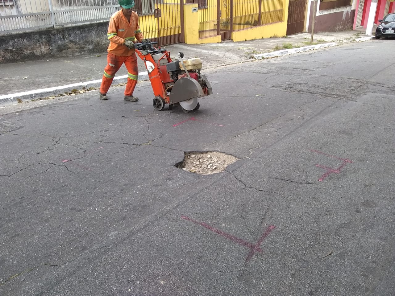 #PraCegoVer - Na primeira foto, trabalhador da Subprefeitura maneja máquina de corte de asfalto, para preparar o piso. Ao fundo, há uma casa toda amarela (paredes e muro). Na segunda foto, o buraco já tapado.