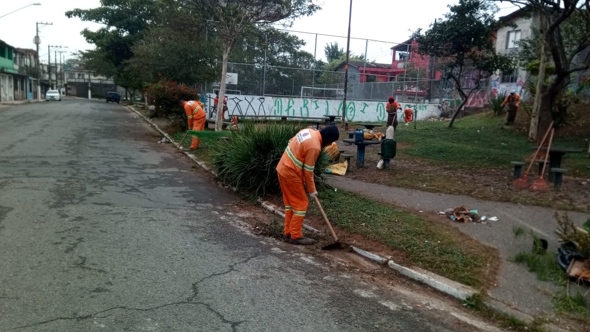 #PraCegoVer - Trabalhadores da Subprefeitura limpam a sarjeta da rua, ao mesmo tempo em que outros cortam a grama do local. Ao fundo, há uma quadra de esportes com alambrado alto.