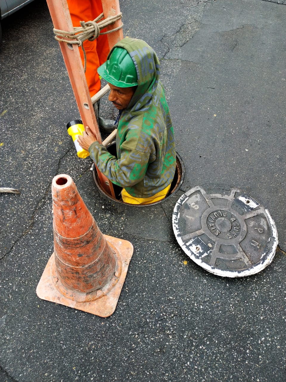 #PraCegoVer - Trabalhador da Subprefeitura desce por uma escada para dentro do poço de visita (PV), ou bueiro, no meio da via. A tampa do bueiro está afastada, de lado. Um cone laranja e branco sinaliza o local.