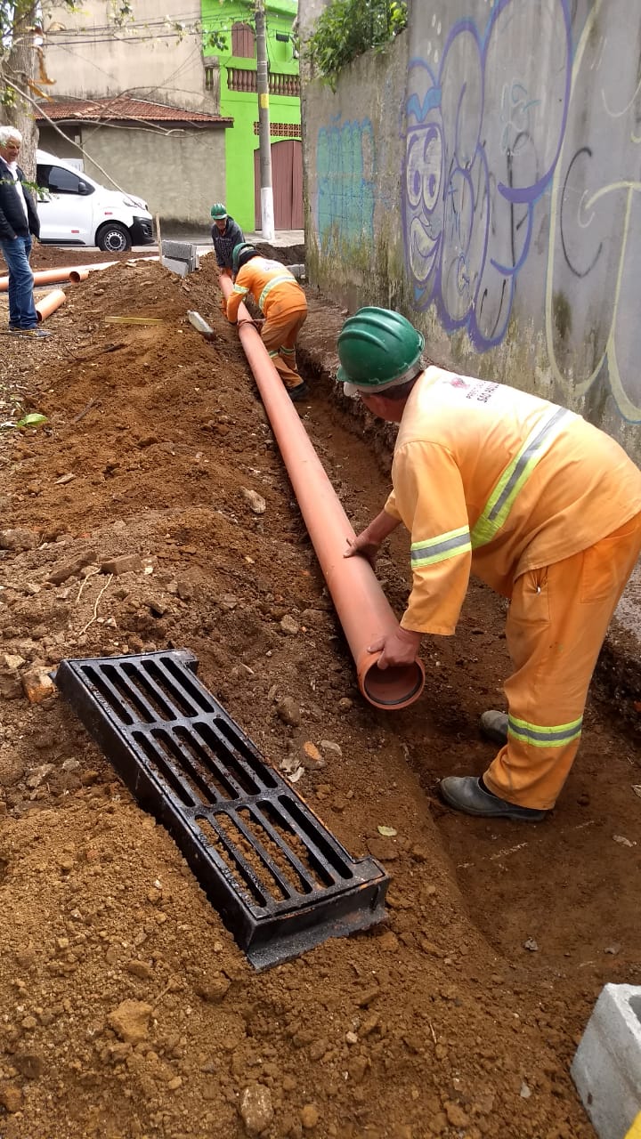 #PraCegoVer - Trabalhadores da Subprefeitura assentam tubos de PVC para o sistema de galeria de águas pluviais (GAP) em buraco previamente cavado. De lado, há uma boca-de-leão (grelha) para captação da água de chuva. Do lado direito, há um muro de uma casa.