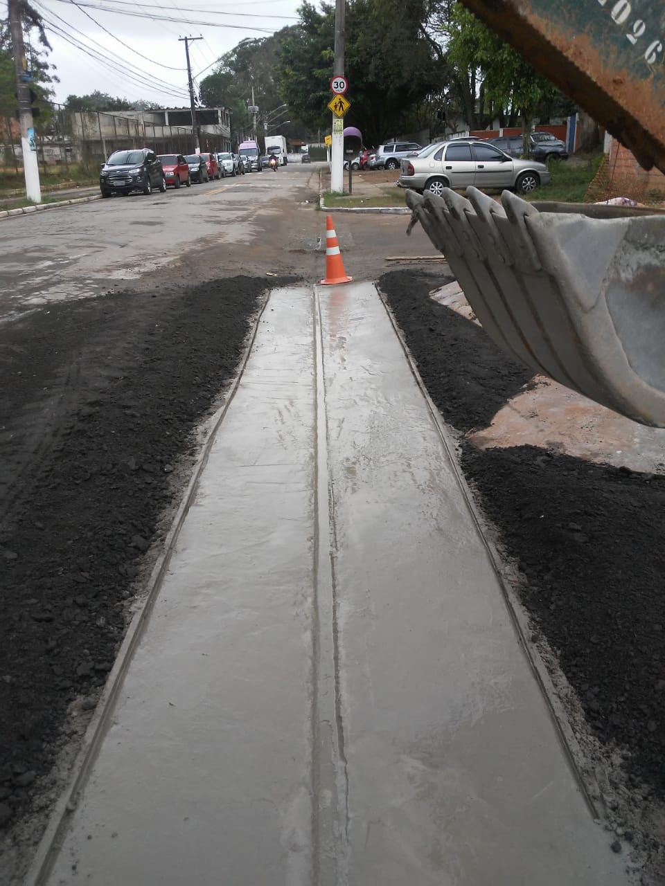 #PraCegoVer - Concreto acabado de colocar no sarjetão da via. O sarjetão é uma sarjeta que atravessa ruas para conduzir as águas pluviais. De cada lado do sarjetão, foi feito asfaltamento.