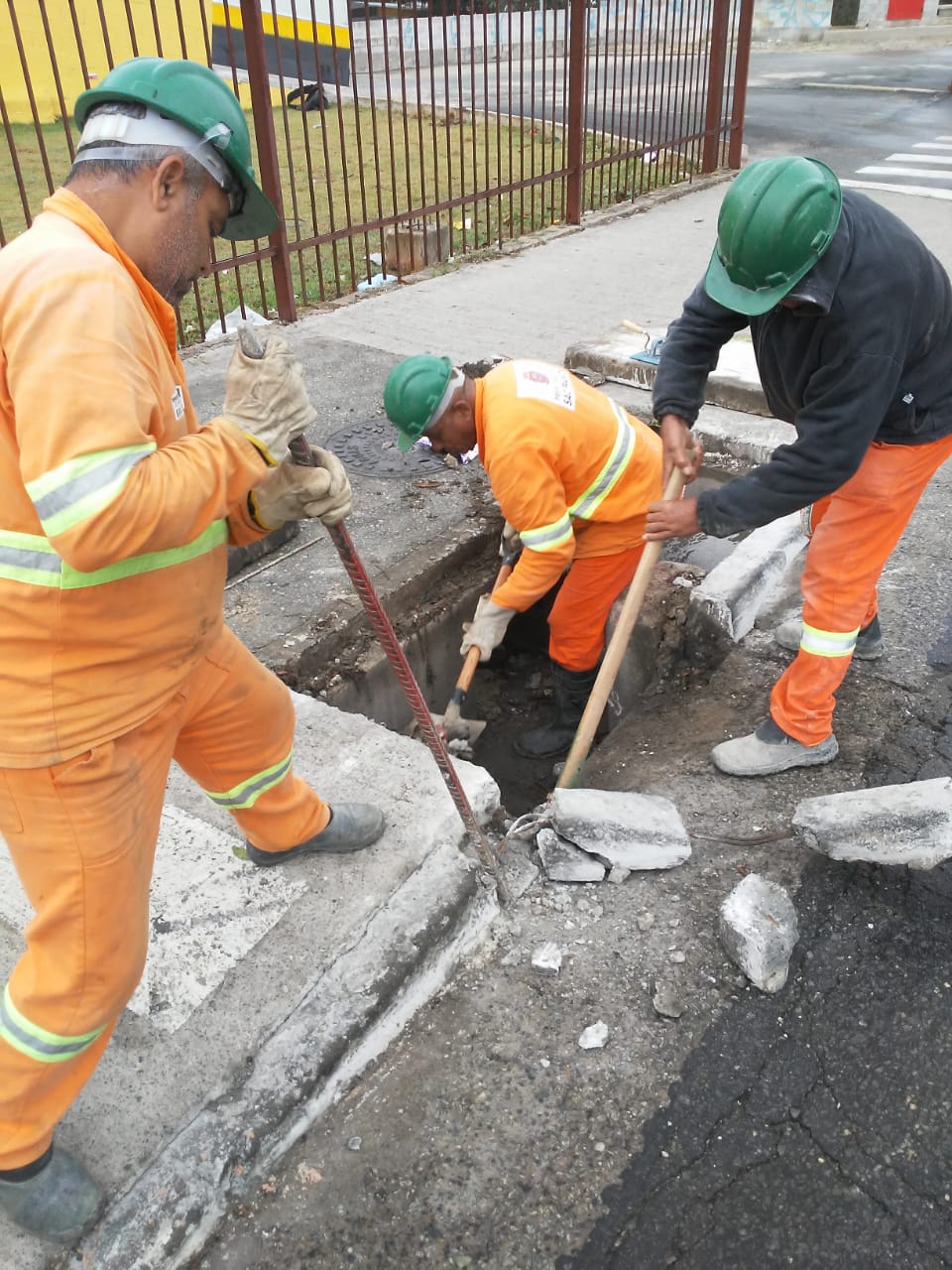 #PraCegoVer - Três trabalhadores da Subprefeitura desmontam a estrutura da boca-de-lobo. Na via, estão pedaços das guias retiradas. Ao fundo, há uma cerca de ferro.
