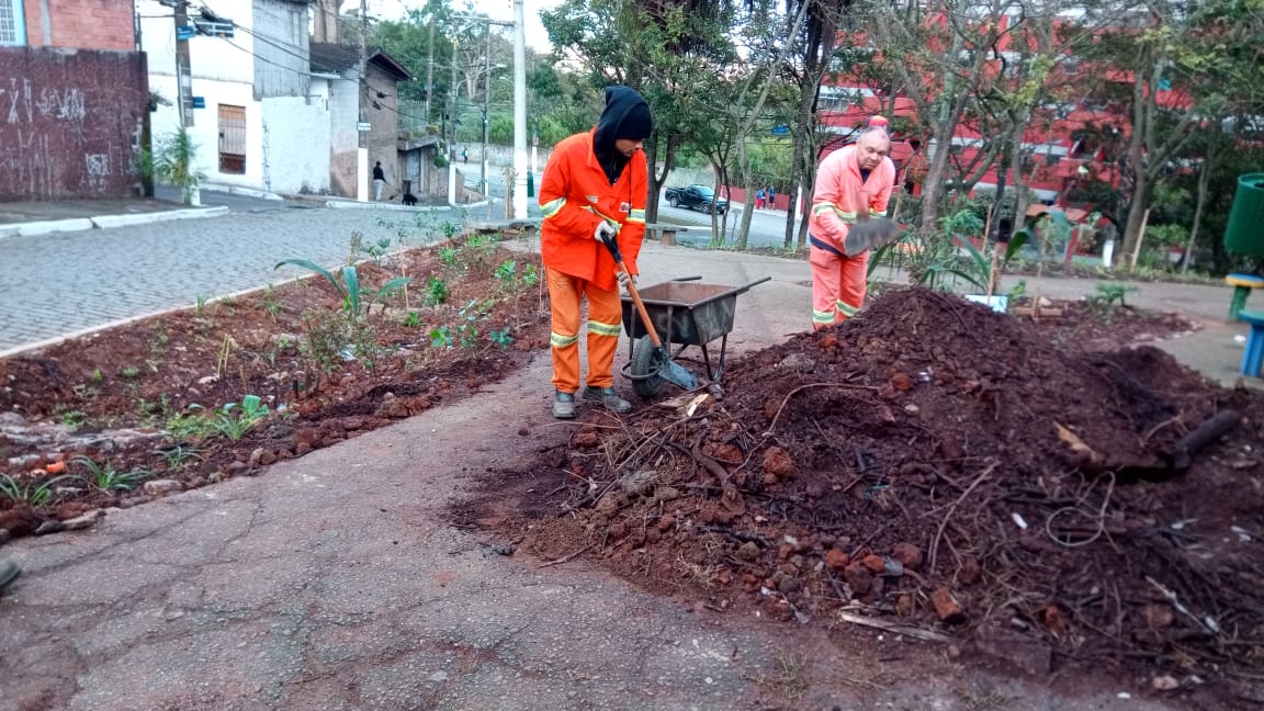 #PraCegoVer - Dois trabalhadores da Subprefeitura movimentam terra nos canteiros do local, uma pequena pracinha na curva da rua. Há um monte de terra do lado direito. E árvores ao fundo.