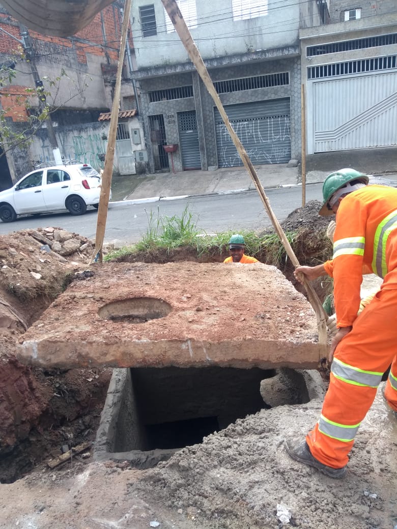 #PraCegoVer - Dois trabalhadores da Subprefeitura supervisionam o levantamento de uma laje por meio de tirantes de cintas de lona. A laje tem um furo à esquerda. Embaixo da laje, há uma caixa coletora de águas pluviais.