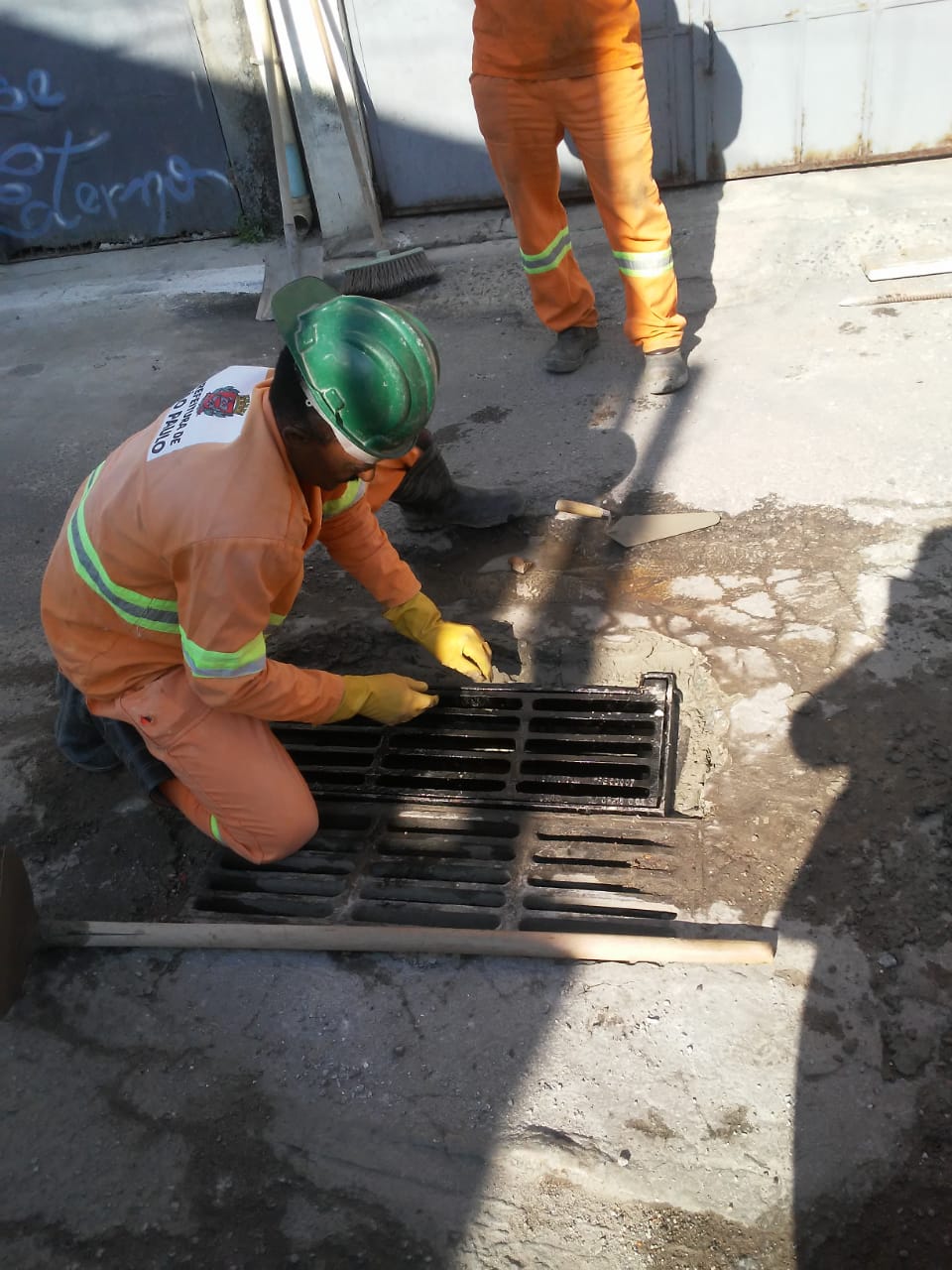 #PraCegoVer - Dois trabalhadores da Subprefeitura instalam grelha de ferro na via.
