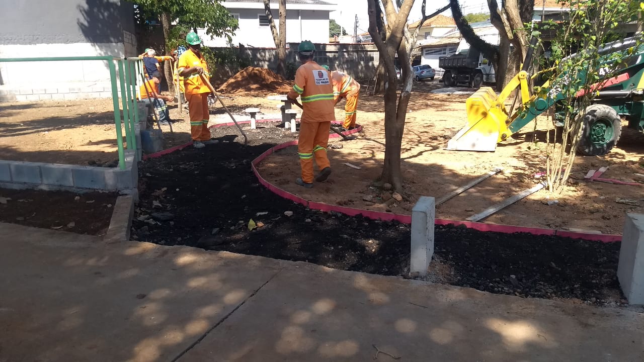 #PraCegoVer - Trabalhadores da Subprefeitura espalham a base para construção de passeio. Há árvores na praça. E uma escavadeira verde e amarela do lado direito.