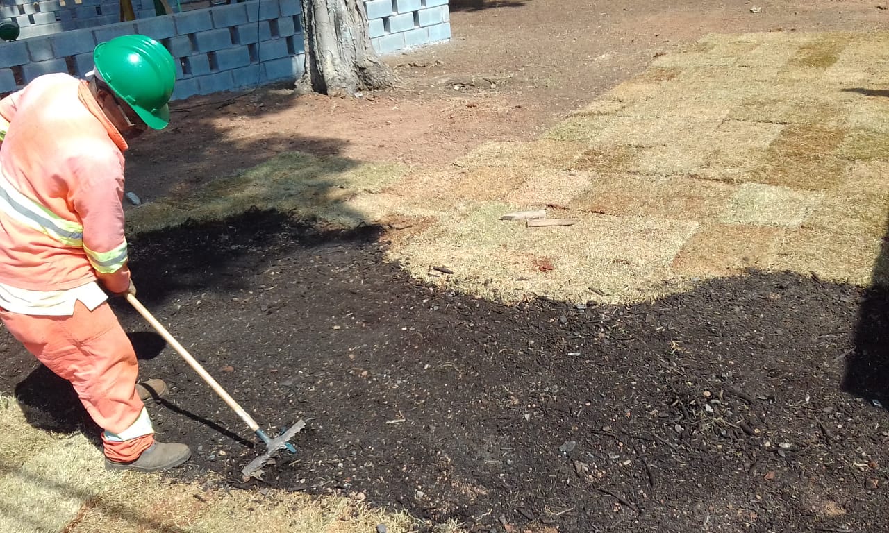 #PraCegoVer - Trabalhador da Subprefeitura espalha terra perto de uma série de tapetes de grama. Ao fundo, há um muro azul.