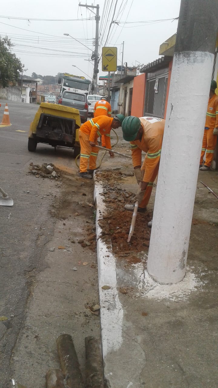 #PraCegoVer - Quatro trabalhadores da Subprefeitura escavam o passeio para rebaixamento da guia. Do lado direito, há um poste. Ao fundo, máquina de britadeira.