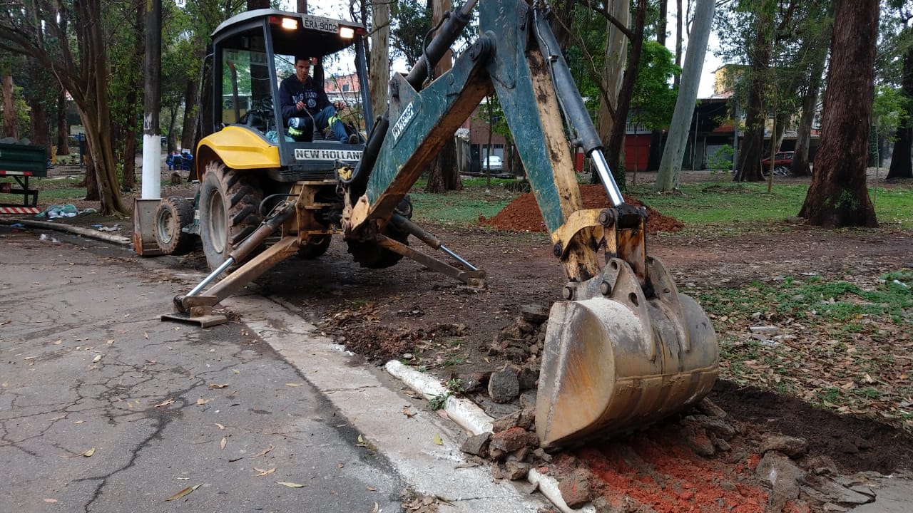#PraCegoVer - Escavadeira remove o passeio atual para proceder ao levantamento das guias. Ao fundo, há árvores situadas na praça.
