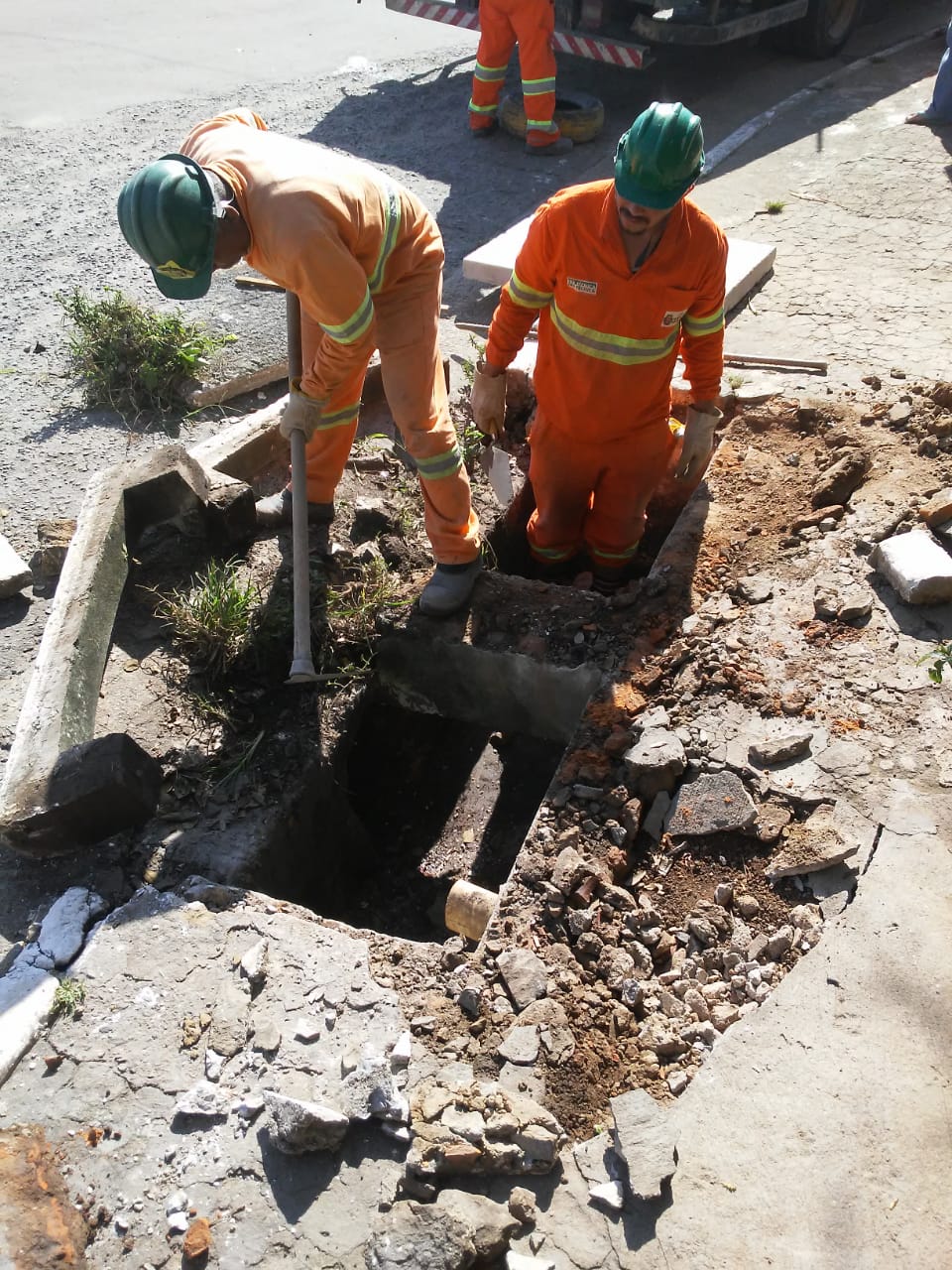 #PraCegoVer - Trabalhadores da Subprefeitura retiram a boca-de-lobo quebrada para instalação de uma nova. Há pedras e pedaços de cimento espalhados pelo chão.