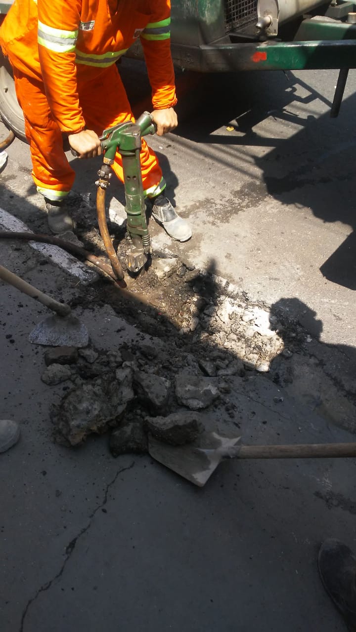 #PraCegoVer - Trabalhador da Subprefeitura usa martelo pneumátioc para destruir a sarjeta quebrada. Ao lado há uma pá.