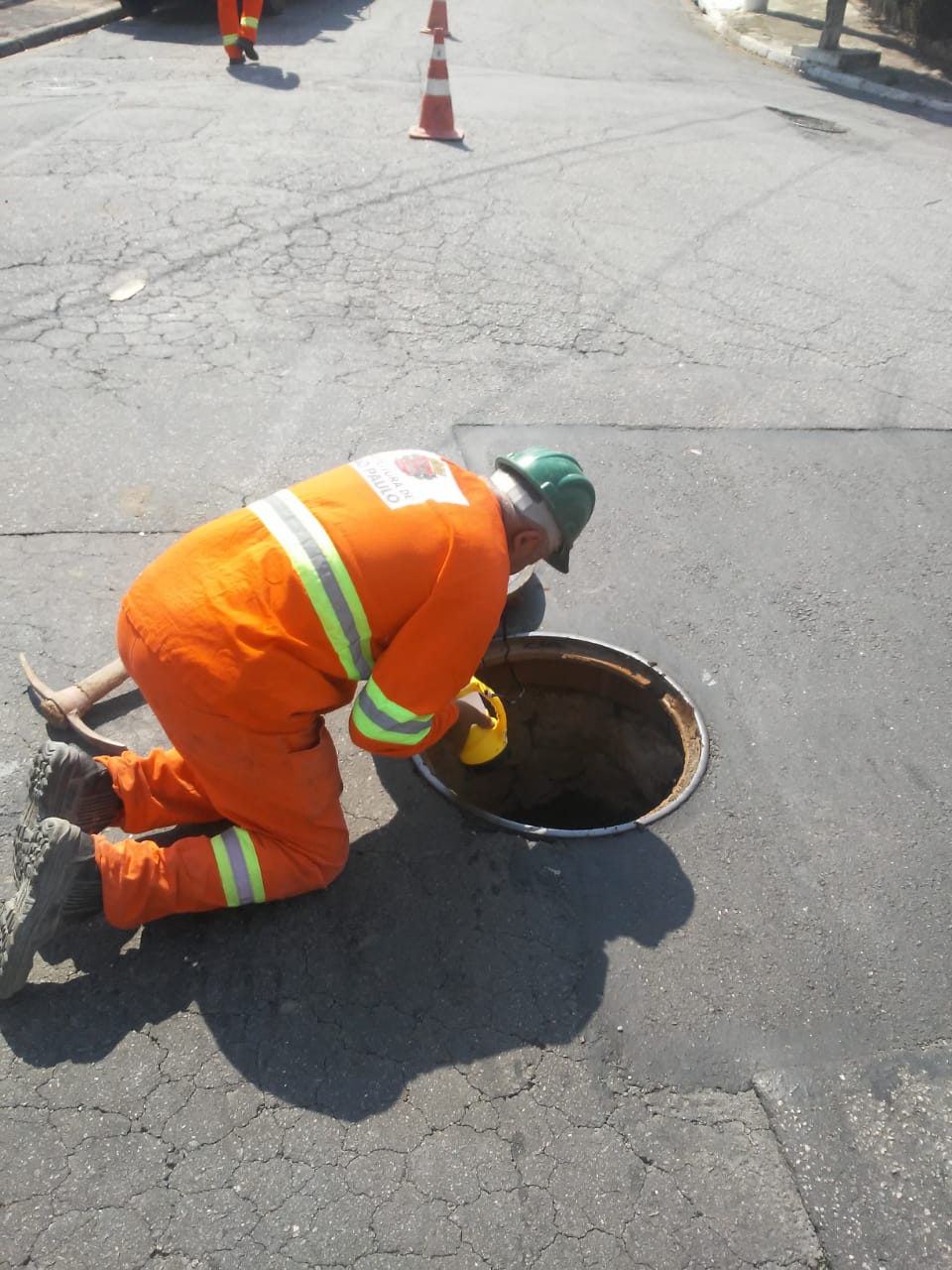 #PraCegoVer - Trabalhador da Subprefeitura inspeciona poço-de-visita na via. O local está sinalizado por cones laranja e branco.