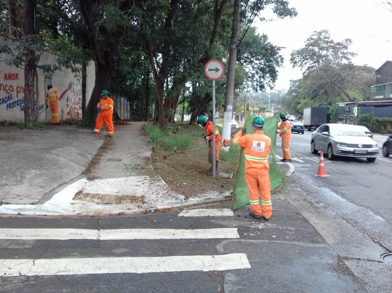 #PraCegoVer - Trabalhadores da Subprefeitura cortam grama na via. Outros dois seguram a proteção de tela.