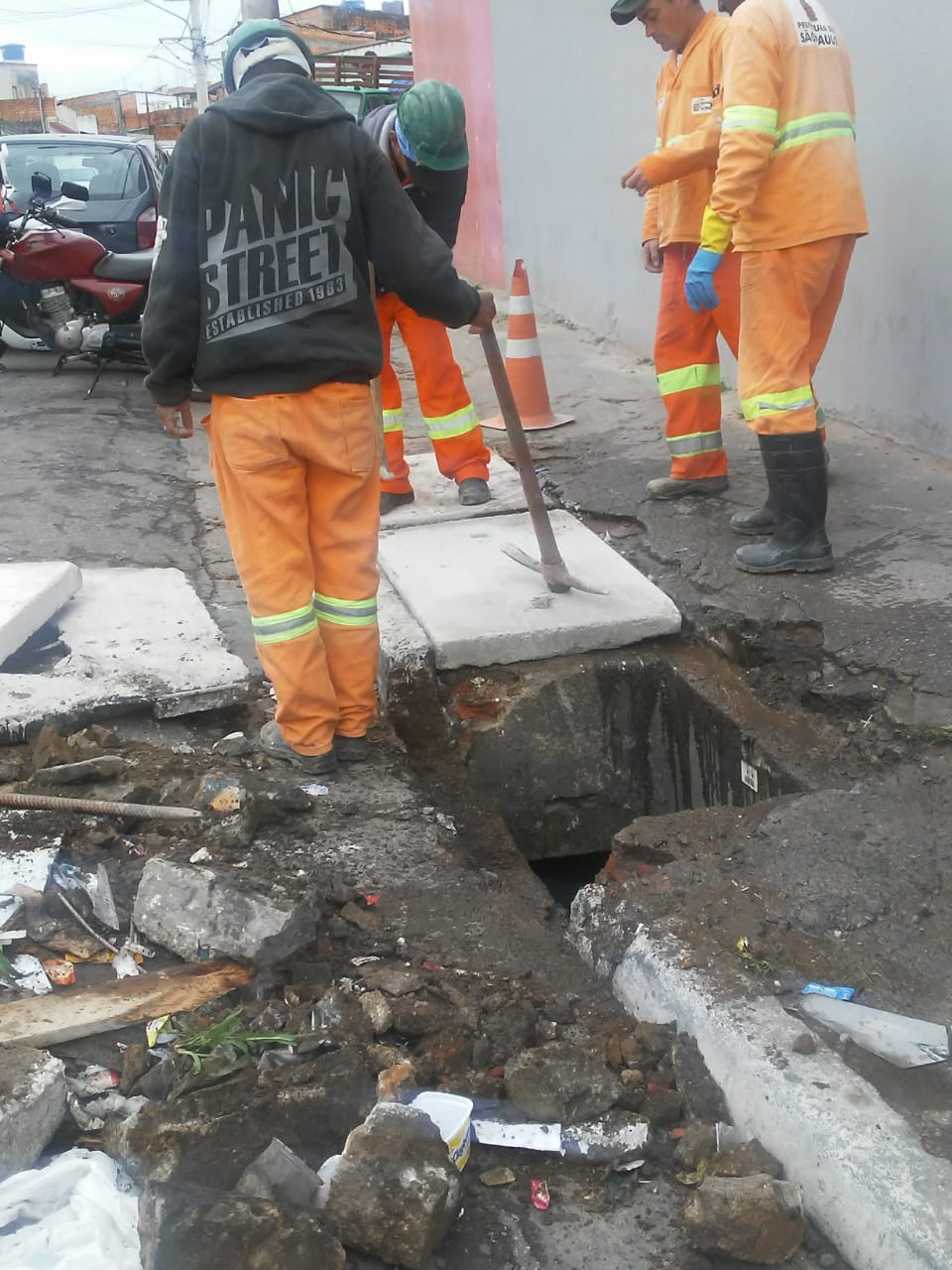 #PraCegoVer - Trabalhadores da Subprefeitura retiram tampa quebrada de boa-de-lobo. Os destroços da tampa quebrada estão espalhados na rua.