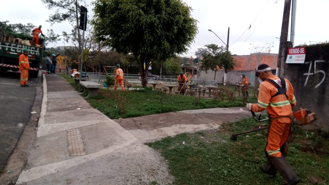 #PraCegoVer - Trabalhadores da Subprefeitura cortam grama em uma pracinha lateral à via. Outros retiram mato do jardim. Há um caminhão da empreiteira contratada do lado esquerdo.