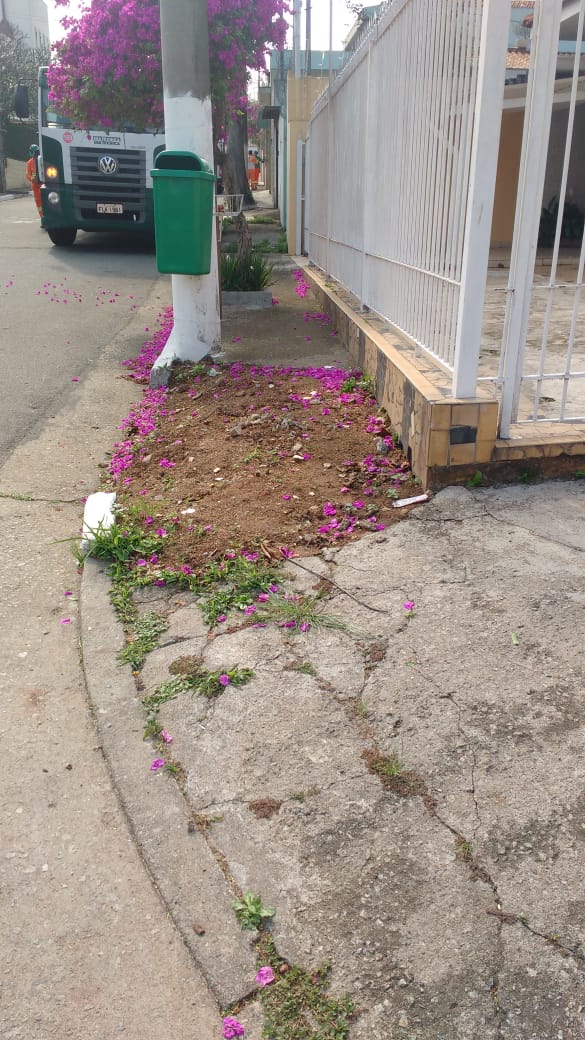 #PraCegoVer - Trecho da calçada perto de uma cerca de ferro. O trecho tem terra aparente e guias quebradas. Em um poste próximo há uma lixeira. Um caminhão da empreiteira está ao fundo.