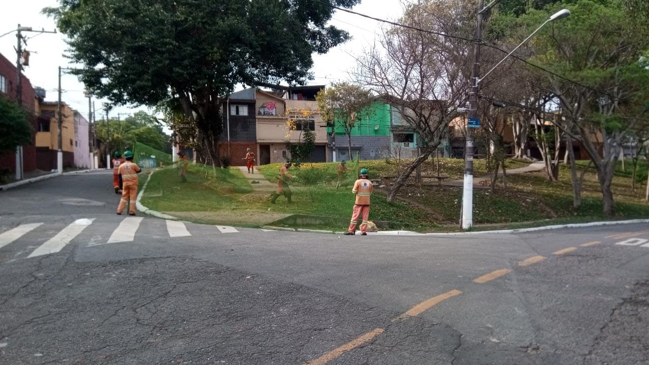 #PraCegoVer - Trabalhadores da Subprefeitura cortam grama com roçadeiras portáteis. Outros seguram a proteção de tela. O local é uma pequena praça à beira da rua.