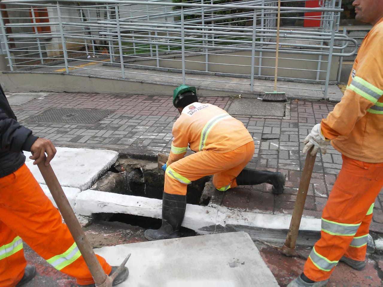 #PraCegoVer - Trabalhadores da Subprefeitura trocam tampa de boca-de-lobo na calçada diante de um banco, onde há acesso em rampa para cadeirantes.