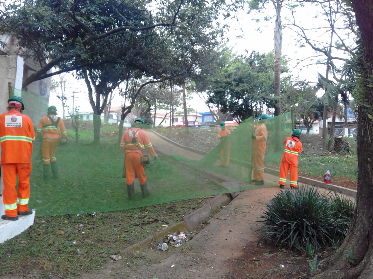 #PraCegoVer - Trabalhadores da Subprefeitura cortam grama na praça, que está sendo reformada. Outros seguram uma proteção de tela. Há um passeio no centro da praça. E árvores.