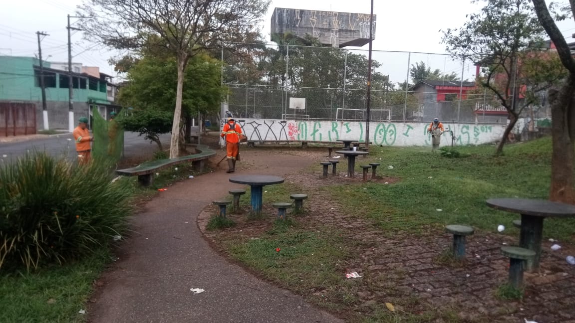 #PraCegoVer - Trabalhadores da Subprefeitura cortam grama em um largo na via. Outros seguram a proteção de tela. Há mesas e bancos de concreto.