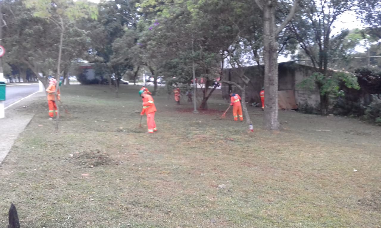 #PraCegoVer - Trabalhadores da Subprefeitura cortam grama em largo na avenida. Outros trabalhadores recolhem a grama cortada.