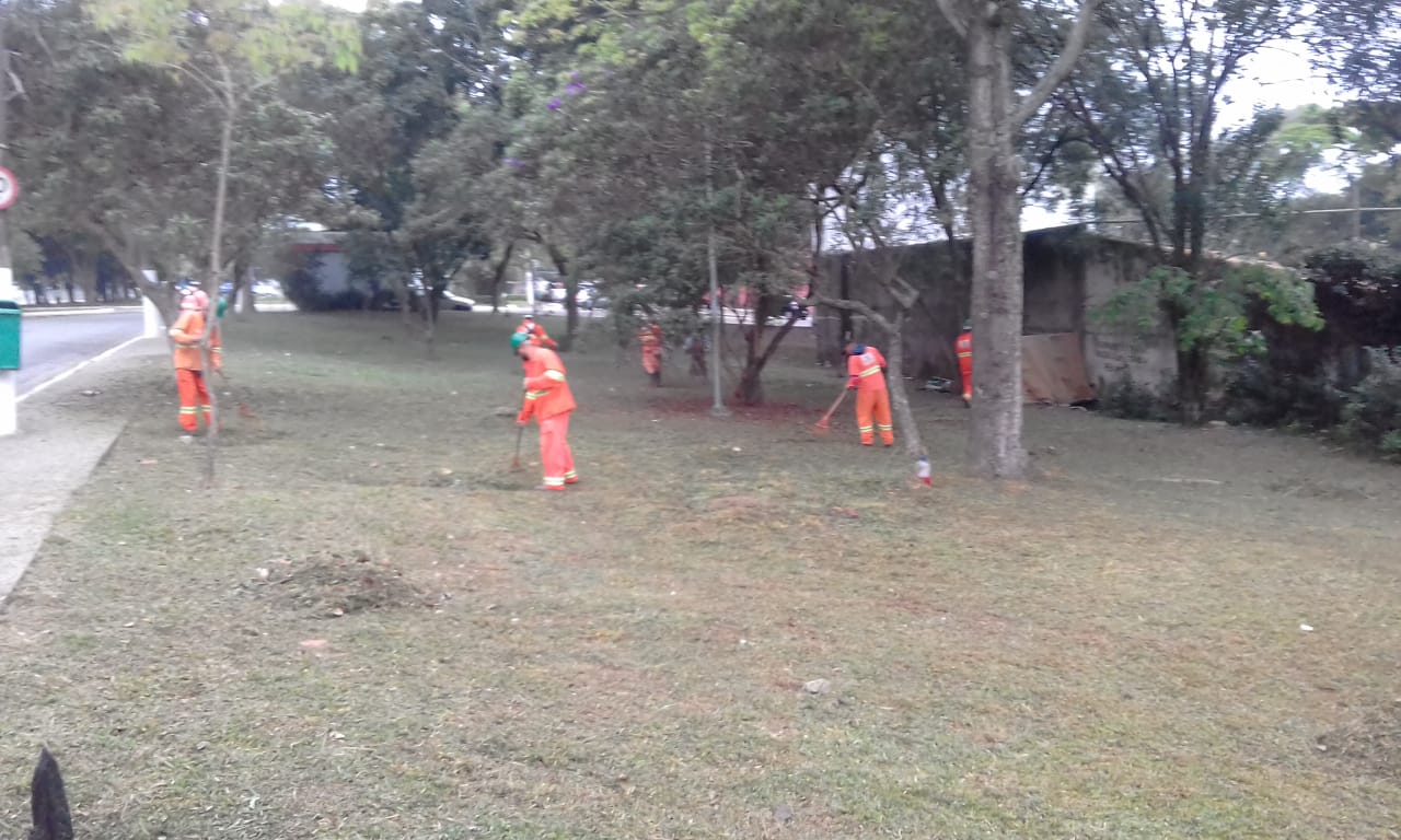 #PraCegoVer - Trabalhadores da Subprefeitura cortam grama em largo na avenida. Outros trabalhadores recolhem a grama cortada.