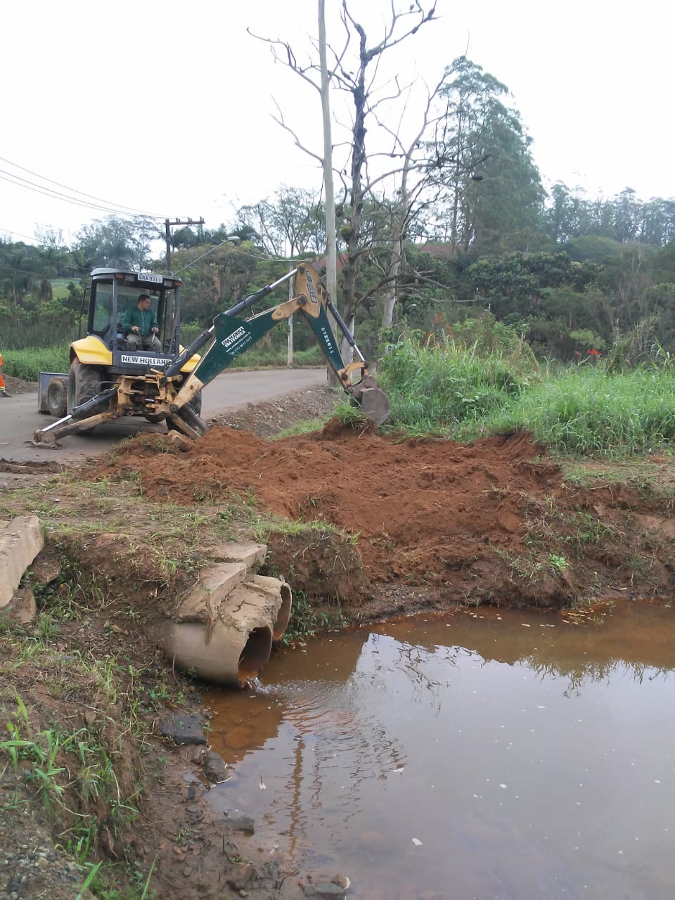 #PraCegoVer - Escavadeira verde e amarela movimenta erra perto de uma travessia sobre tubos de concreto.