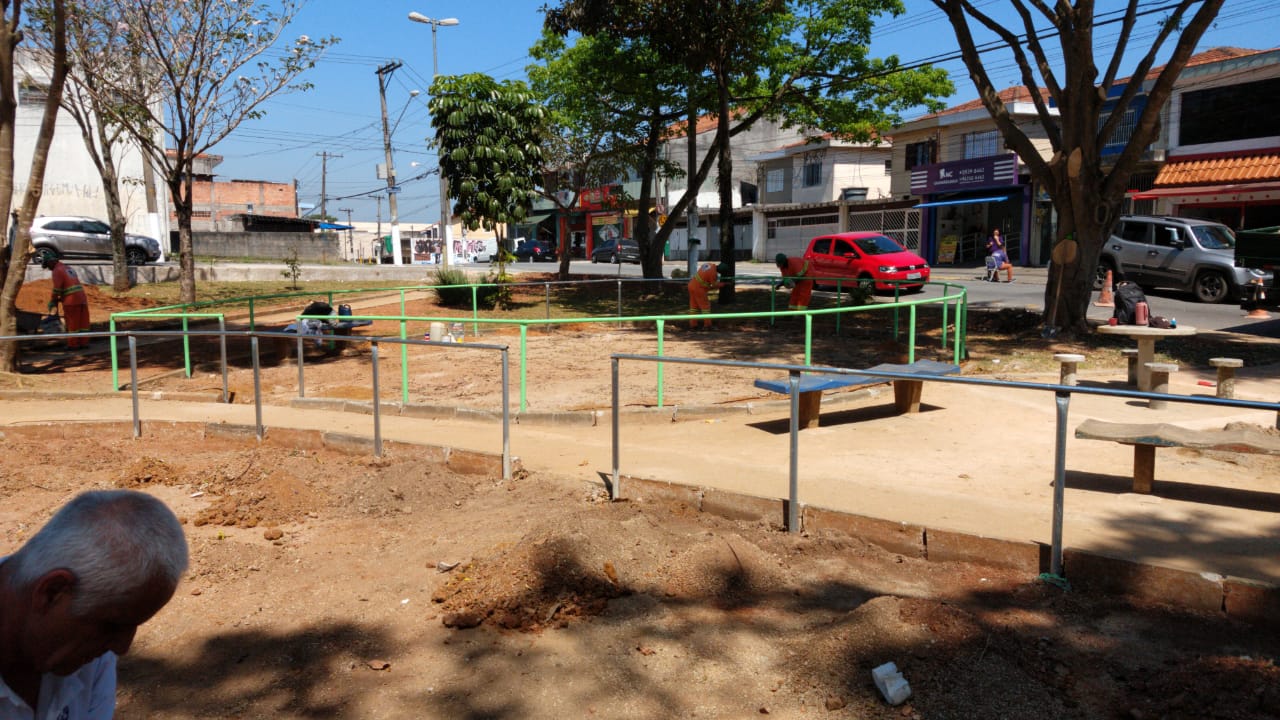 #PraCegoVer - Instalação de cercas de metal na praça. Ao fundo, passam carros. Na praça, há árvores.