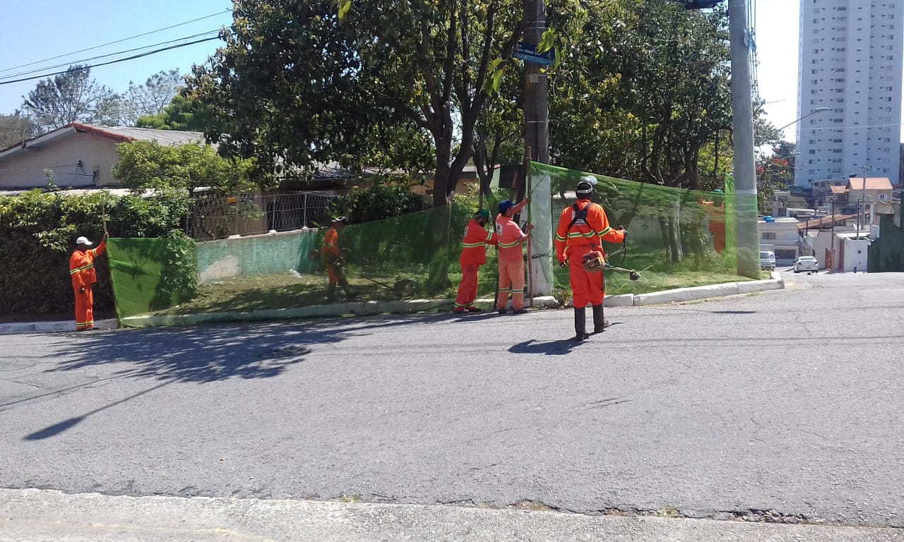 #PraCegoVer - Trabalhadores da Subprefeitura cortam grama na via, que é uma esquina, com árvores na calçada. Outros trabalhadores seguram proteções de tela.