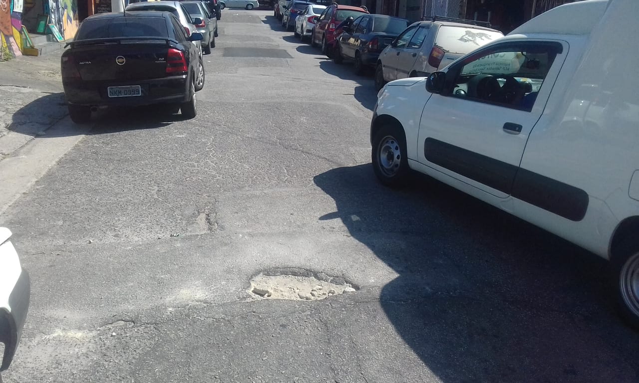 #PraCegoVer - Na primeira foto, está o buraco original. Passa um carro branco ao lado na rua onde há carros estacionados nos dois lados. Na segunda foto, o remendo já executado.