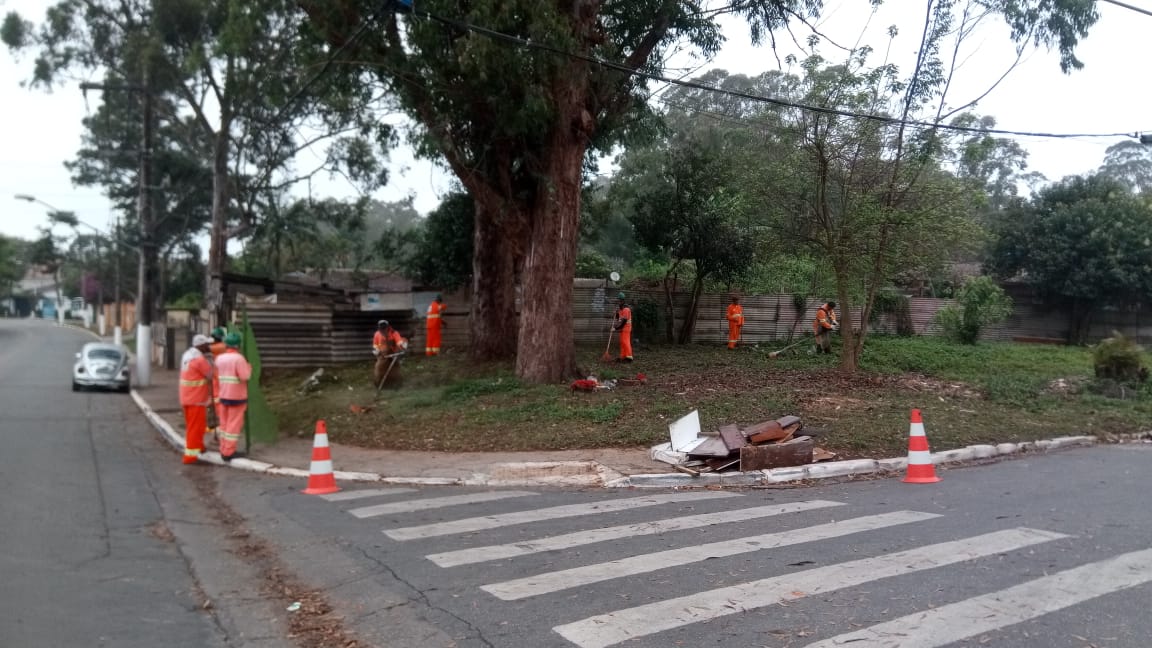 #PraCegoVer - Trabalhadores da Subprefeitura cortam a grama e limpam o mato. Outros trabalhadores seguram a tela de proteção. Há uma árvore de grande porte no centro.